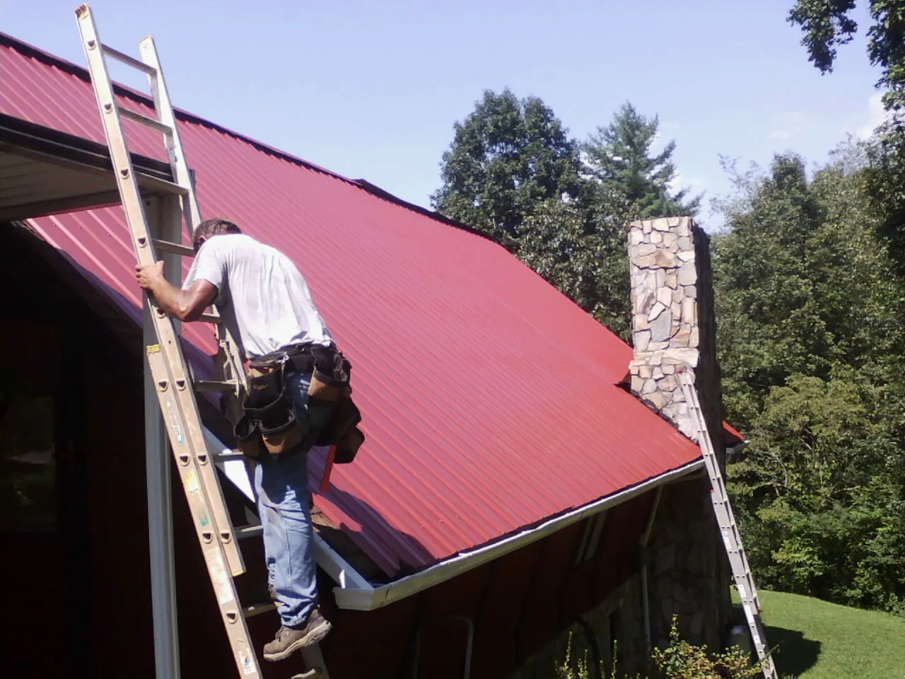 Roofer on a ladder beside a red metal roof and stone chimney — Soffit & Fascia Repair by Roofing Brothers in Grand Ridge